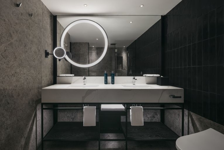 A modern bathroom with dual sinks and a large, circular mirror illuminated from behind. Dark tiles cover the walls, contrasting with the white countertop and towels.