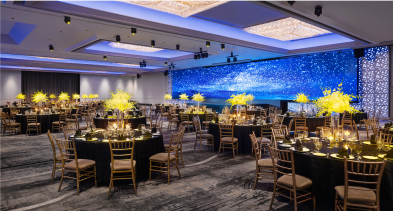 Round tables with yellow centerpieces set for an event in a room with blue illuminated walls and star-like lights.