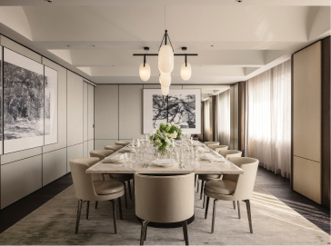 A modern dining room features an elongated table set for a meal, surrounded by upholstered chairs, with artistic monochrome wall panels, pendant lights, and sheer draped windows.