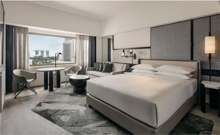 A neat, modern hotel room with a large bed centered between two nightstands and lights, a sitting area by the window offering a city view, and an overall elegant decor.