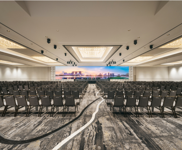 Rows of empty chairs face a large screen displaying a cityscape within a spacious conference room with patterned carpeting.