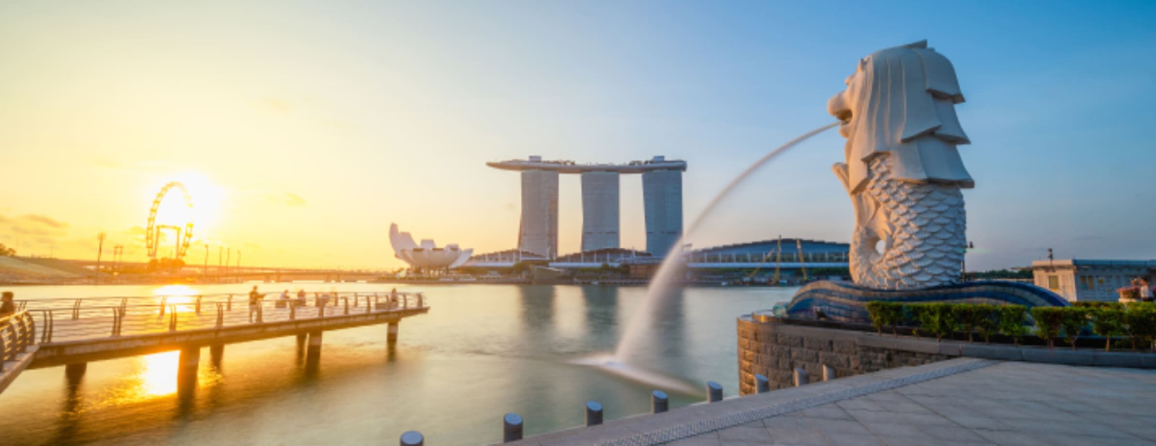 A large sculpture with a fountain jet streams water against the backdrop of a sunset, with a city skyline featuring a distinctive three-towered building and a Ferris wheel.