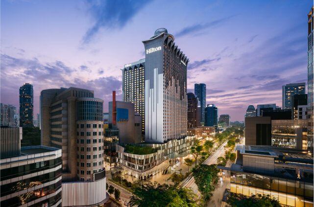 A towering hotel at dusk dominates the skyline amidst a backdrop of city buildings, illuminated under a fading sky.