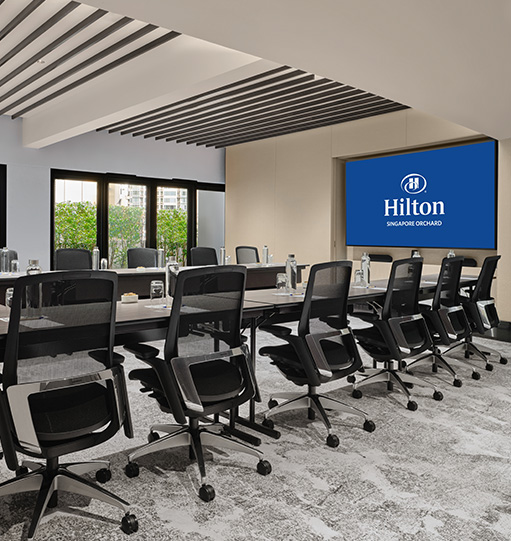 A modern conference room with a large table and chairs facing a screen displaying the Hilton logo and 'SINGAPORE ORCHARD.' Sunlight streams in through windows with a city view.